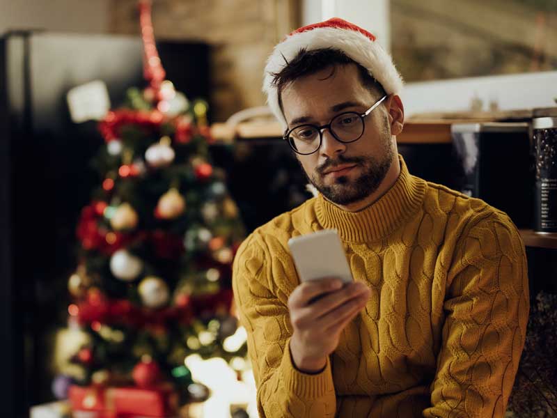 A man in a yellow sweater and santa hat texts on a cellphone in front of a christmas tree