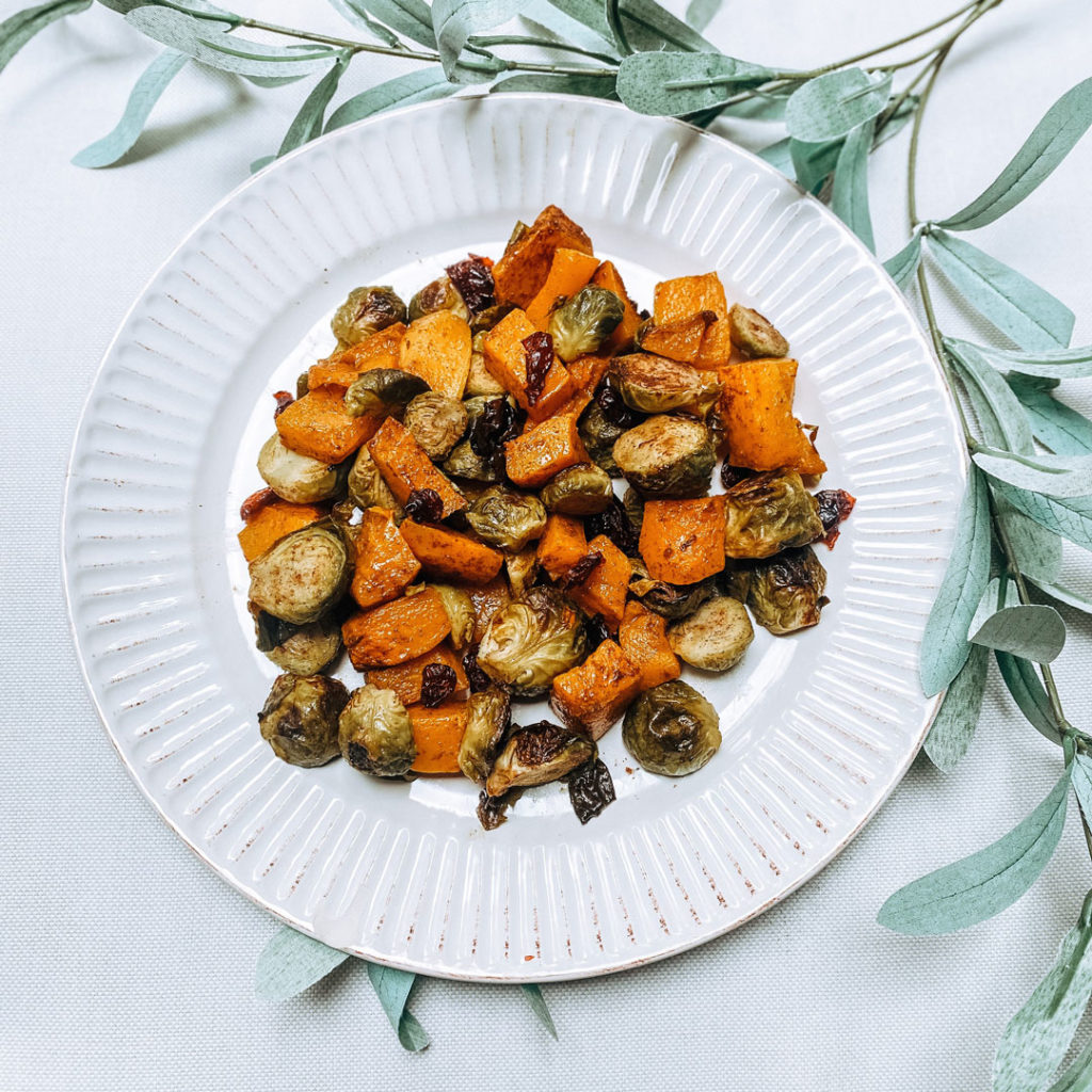 An overhead view of roasted brussel sprouts and squash on a white plate, white table cloth with greenery for decoration
