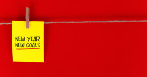 a red background with a string stretched horizontally. A yellow note with the words "New Year. New Goal" is pinned to the string.