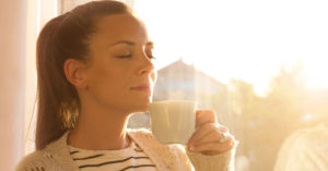 a woman takes a deep breath to smell the fresh cup of coffee in her mug while near a window with plentiful sun