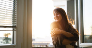 a smiling woman embraces herself near a sunny window