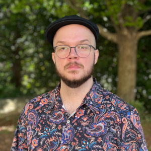 A portrait photo of Colby W. A caucasian man wearing glasses, a newsboy hat, and a colorful paisley shirt.