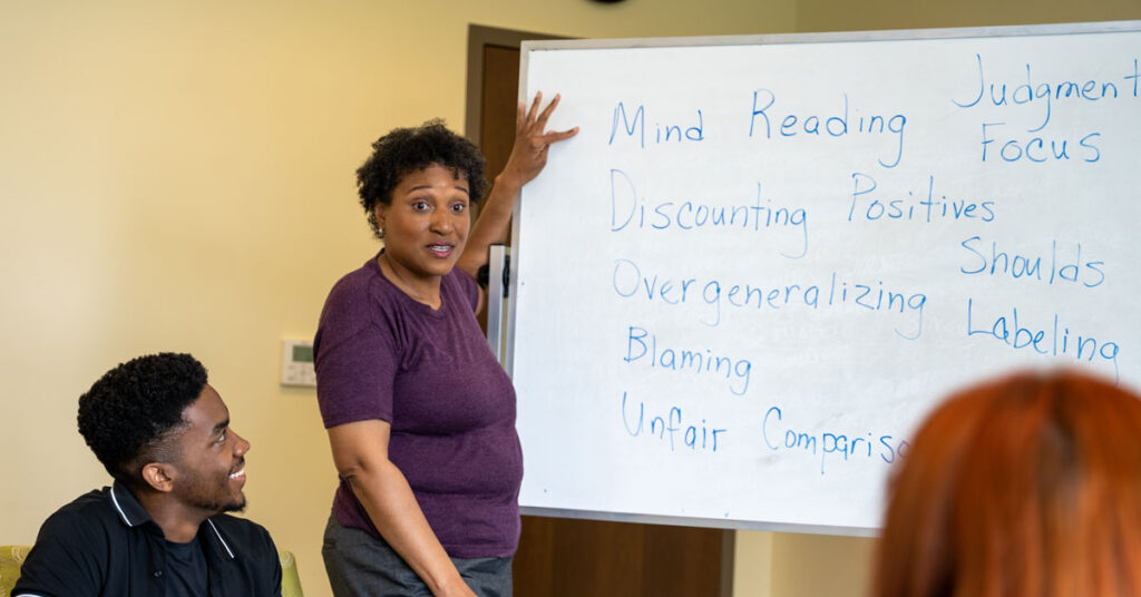 A mental health professional speaks to a group of patients while standing next to a whiteboard filled with examples of cognitive distortions