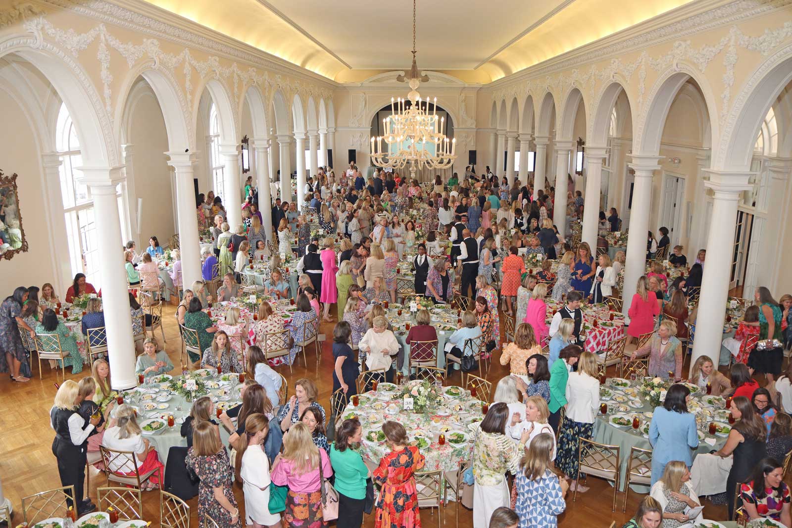 Aerial photo looking down from balcony at ballroom set for a luncheon with hundreds of women conversing and finding their seats