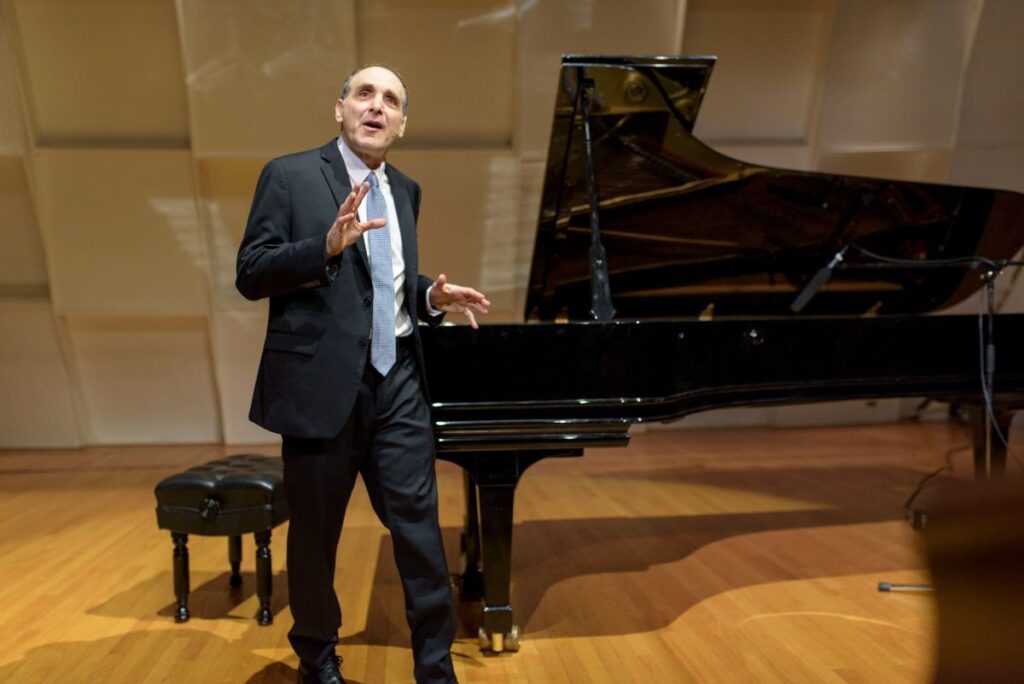 Dr. Kogan stands on stage in front of grand piano delivering a lecture
