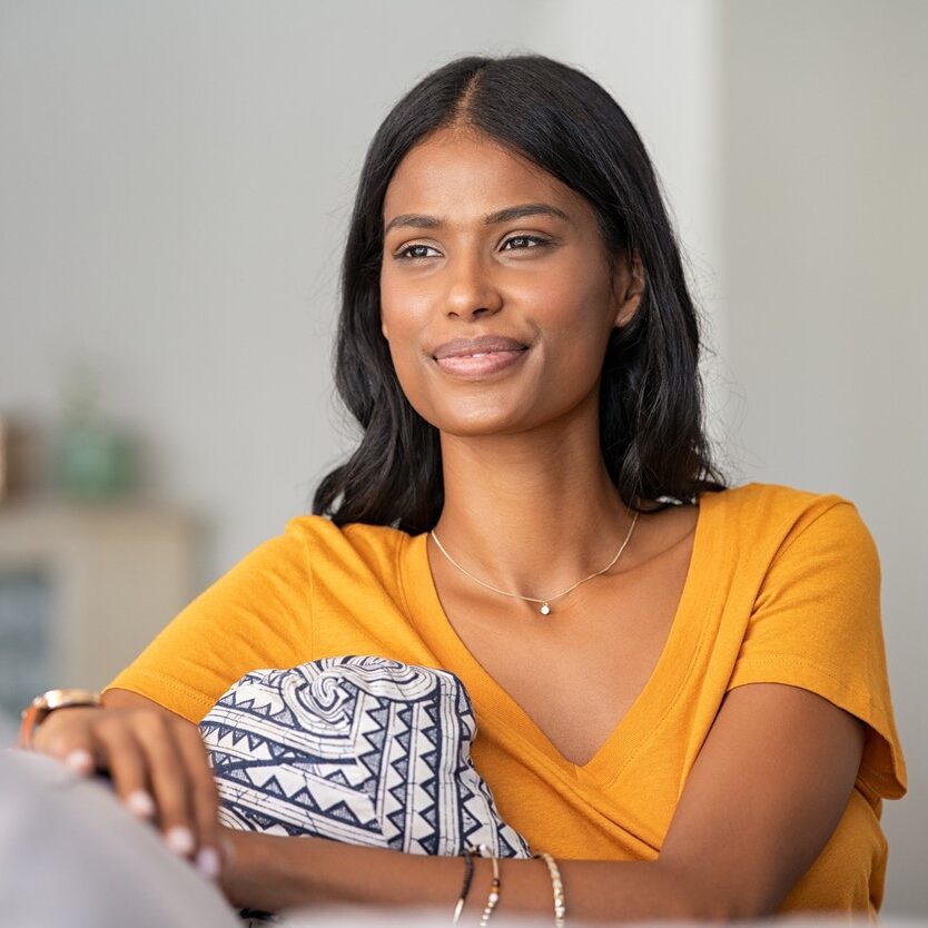 Smiling indian girl relaxing at home Young woman relaxing on couch at home while looking away. Thoughtful smiling woman daydreaming while sitting on sofa at home.
